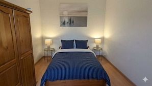 A bedroom with a bed, two bedside tables, a nightstand, a wooden wardrobe, and a wall-mounted picture of a sunset over water. - Duplex near the beach (Les Sables-d'Olonne)