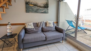 A grey sofa with striped pillows, a painting of a beach scene, and a view of a balcony with a blue chair. - Duplex near the beach (Les Sables-d'Olonne)