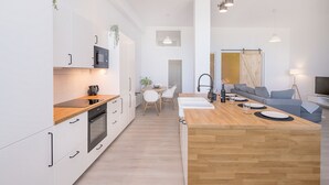 A modern kitchen with white cabinets, a wooden countertop, and a central island. There is a dining area with a table and chairs, and a living area with a sofa and a TV. - Homey Apt. Cómpeta (Málaga)
