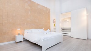 A modern bedroom with a wooden accent wall, a white bed, a nightstand with a lamp, and a walk-in closet. - Homey Apt. Cómpeta (Málaga)