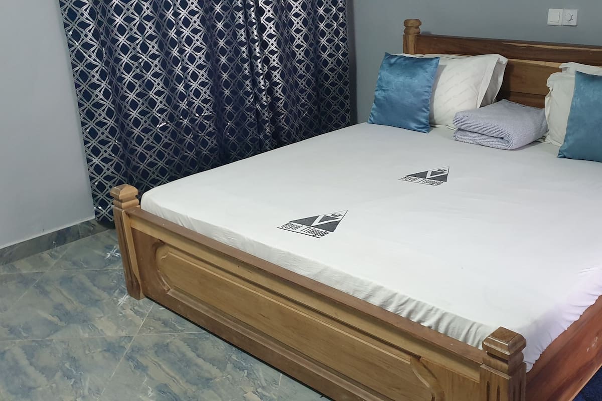 A neatly made bed with a white bedspread and two pillows, one blue and one white, in a room with patterned curtains and a wooden headboard.