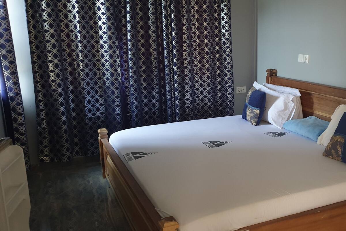 A neatly made bed with a wooden headboard, a patterned curtain, and a plain wall with a light switch.