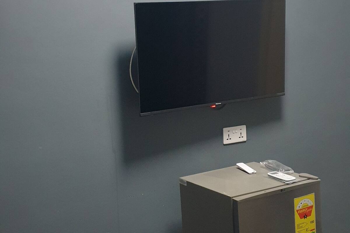 A room with a grey wall, a mounted flat-screen TV, a small refrigerator, and an air conditioning unit.