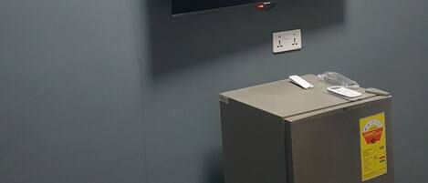 A room with a grey wall, a mounted flat-screen TV, a small refrigerator, and an air conditioning unit.