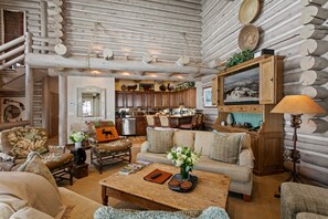 A log cabin interior with a living room, kitchen, and dining area, featuring wooden walls, a fireplace, and rustic decor.