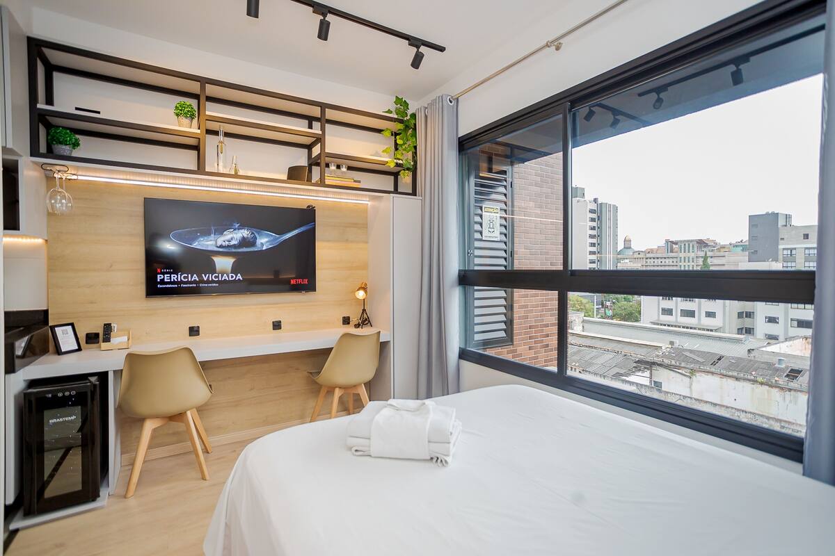 A hotel room with a bed, a desk with a TV, a minibar, and a large window with a city view.