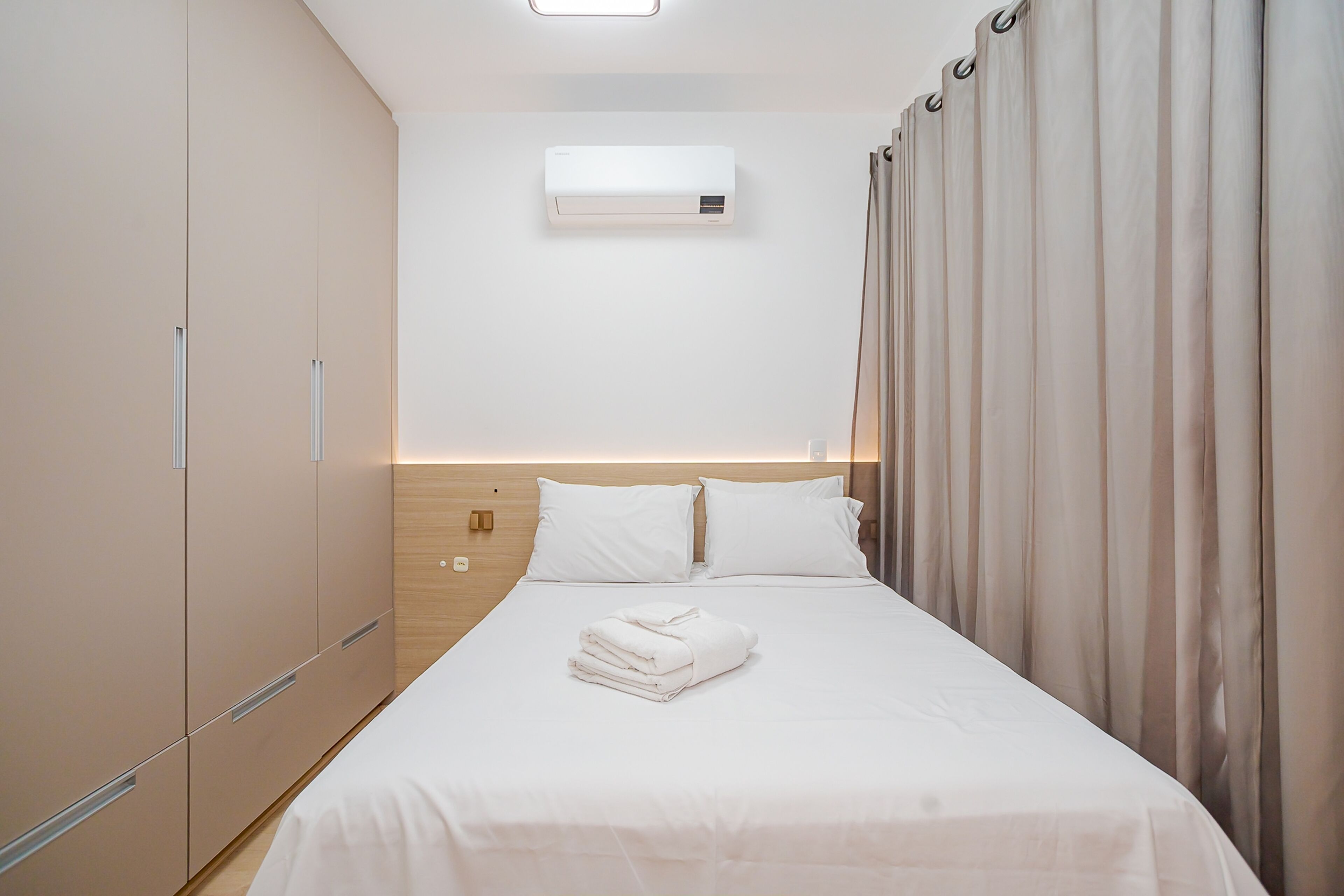 Una cama bien hecha con sábanas blancas y una toalla doblada, un aire acondicionado de pared y un cabecero de madera.