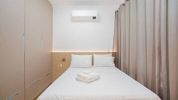 Una cama bien hecha con sábanas blancas y una toalla doblada, un aire acondicionado de pared y un cabecero de madera.