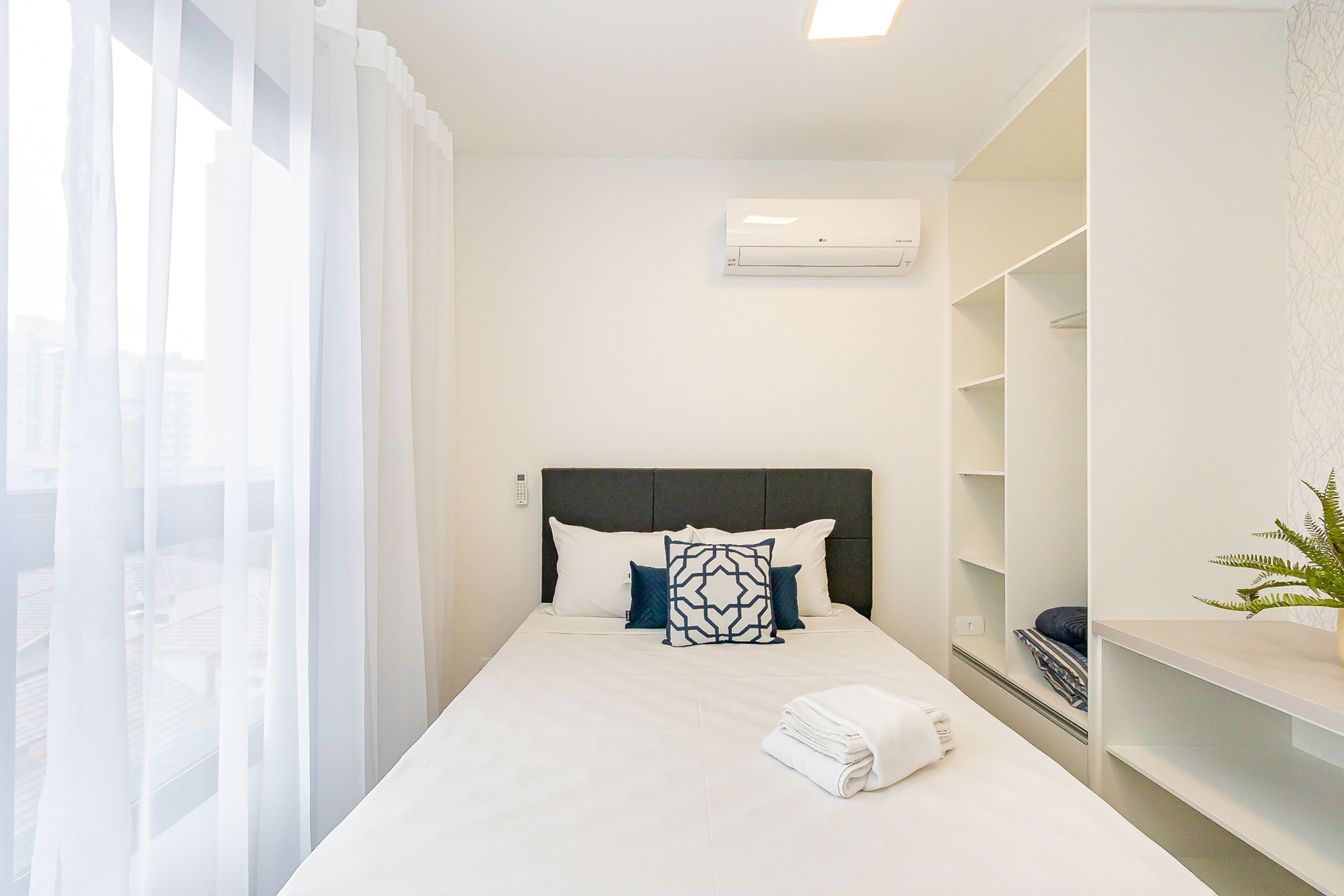 Habitación de hotel con cama, cortinas blancas, cabecera, unidad de aire acondicionado y una planta.