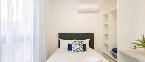 Habitación de hotel con cama, cortinas blancas, cabecera, unidad de aire acondicionado y una planta.