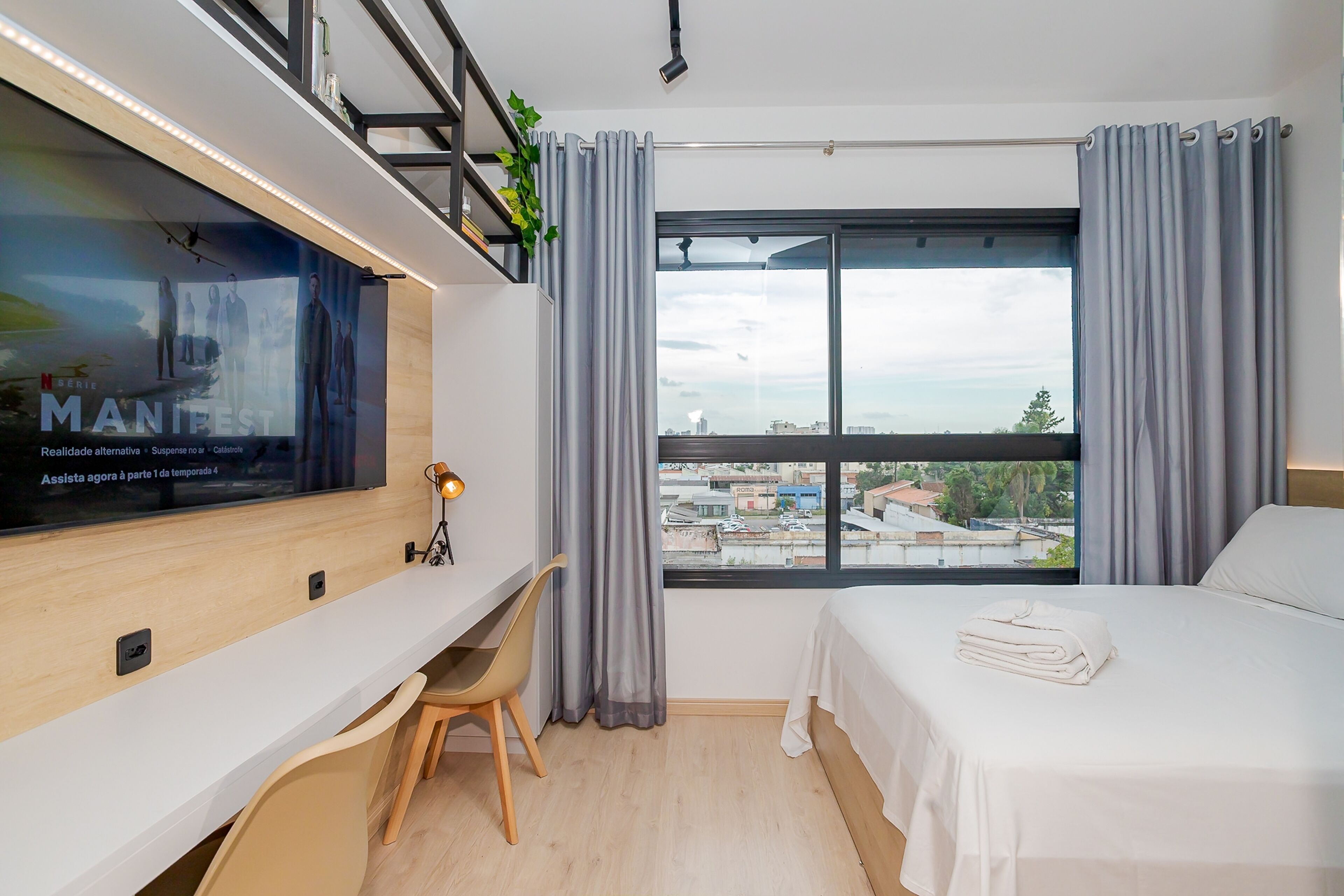 Una habitación de hotel con cama, escritorio, silla, televisor y ventana con cortinas.