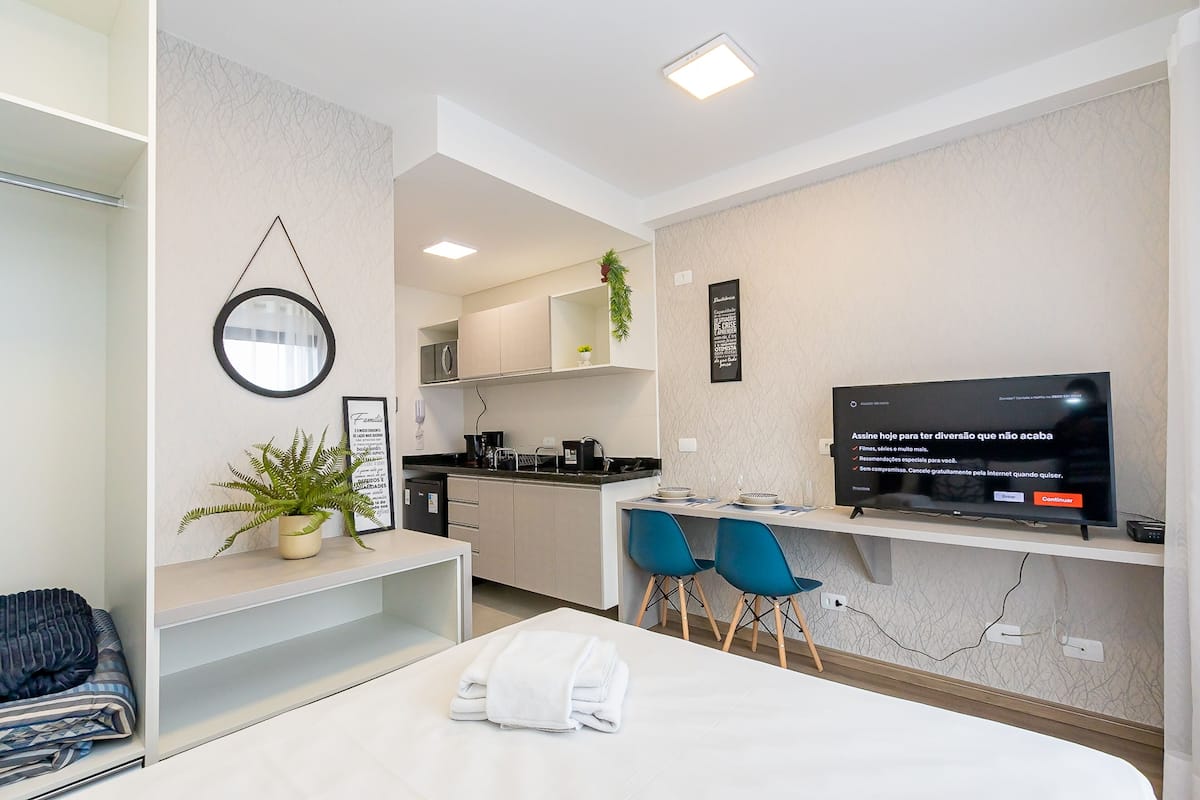 A modern hotel room with a bed, a desk, a TV, a small kitchenette, and a bathroom.