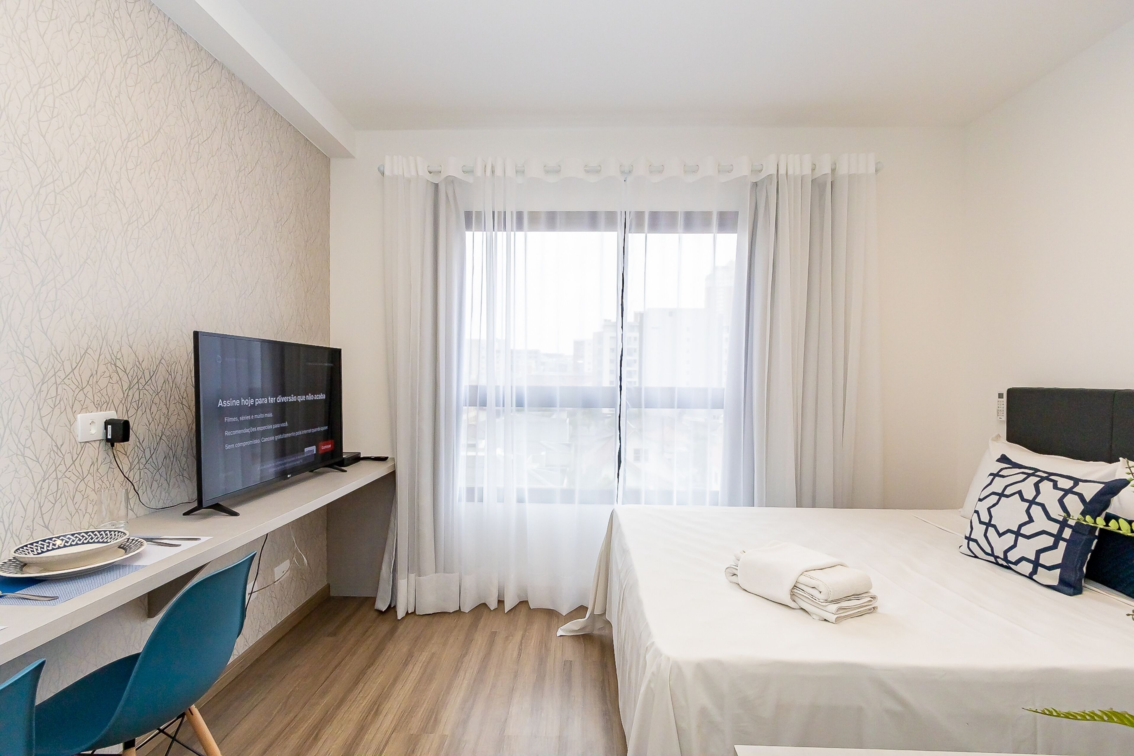 Une chambre d’hôtel avec un lit, un téléviseur posé sur un bureau, une fenêtre avec des rideaux légers et une vue sur la ville.
