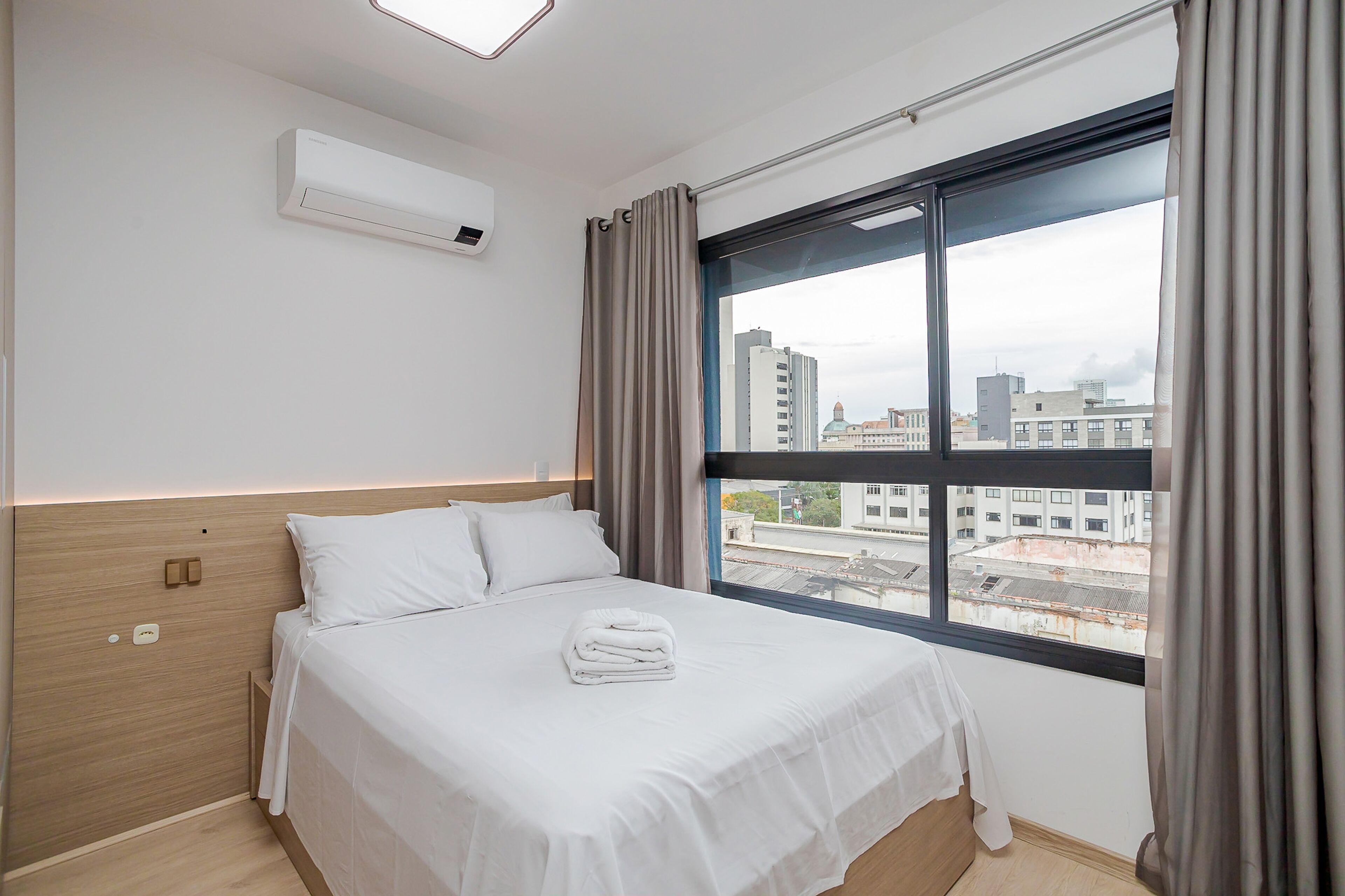 Habitación de hotel con un ventanal grande, cama con ropa blanca, cabecero de madera y un equipo de aire acondicionado en la pared.
