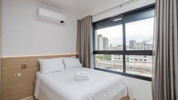 Habitación de hotel con un ventanal grande, cama con ropa blanca, cabecero de madera y un equipo de aire acondicionado en la pared.