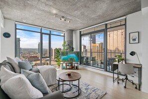 A spacious living room with a large window, a sofa, a coffee table, and a desk with a chair. - DT Views |King Bed |Mins to Stampede | UG Parking (Calgary)