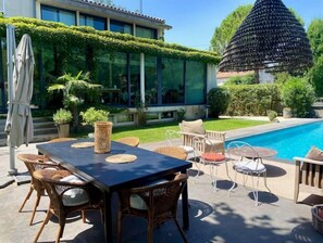 A poolside area with a dining table and chairs, surrounded by greenery and a modern building. - Magnifique Villa Avec Piscine (Arles)