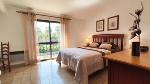 Een slaapkamer met een groot bed, een bureau, een stoel en uitzicht op palmbomen door het raam.