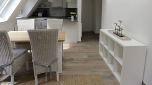 A modern kitchen with white cabinetry, a dining area with wicker chairs, and a white shelving unit. - Vacation apartment Odde (Nebel)