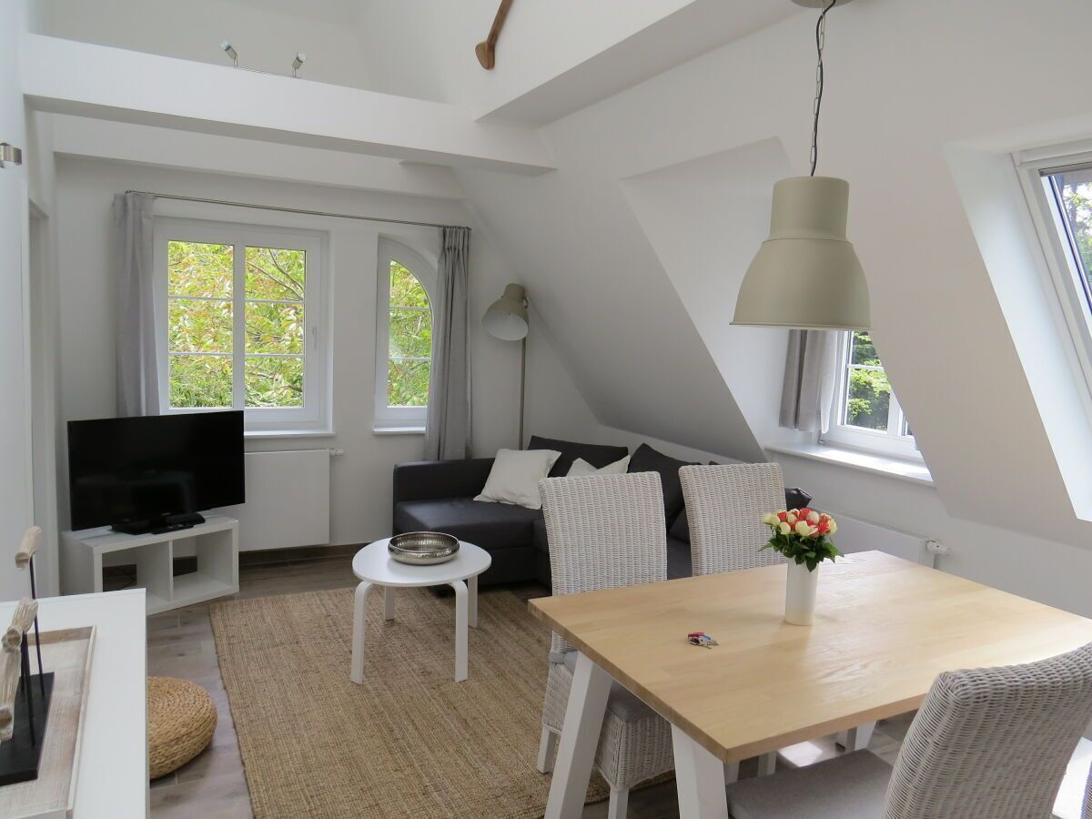 A modern loft-style living space with a dining area, a sofa set, a flat-screen TV, and large windows offering a view of greenery.