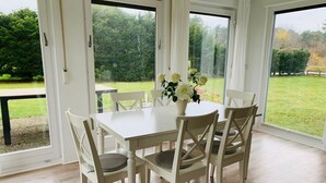 A dining area with a white table and chairs, a vase with flowers, and large windows offering a view of a green garden. - Diese Ferienwohnung Liegt im Erdgeschoss Eines Wunderschön Gelegenen Hauses mit Riesigem Grundstück in Nebel (Nebel)
