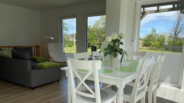 A dining area with a white table and chairs, a grey sofa, and a view of the outdoors through large windows. - Diese Ferienwohnung Liegt im Erdgeschoss Eines Wunderschön Gelegenen Hauses mit Riesigem Grundstück in Nebel (Nebel)