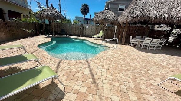 A swimming pool with a brick patio, lounge chairs, and thatched umbrellas.