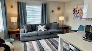 A modern living room with a grey sofa, two side tables with lamps, a coffee maker, a colorful painting, and a striped rug.