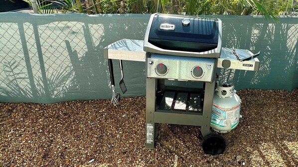 A portable Weber Spirit II grill with a propane tank, set on a gravel surface with a green fence in the background.