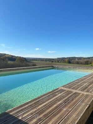 Una piscina cristalina con un muelle de madera en primer plano y una vista panorámica de colinas y árboles al fondo.
