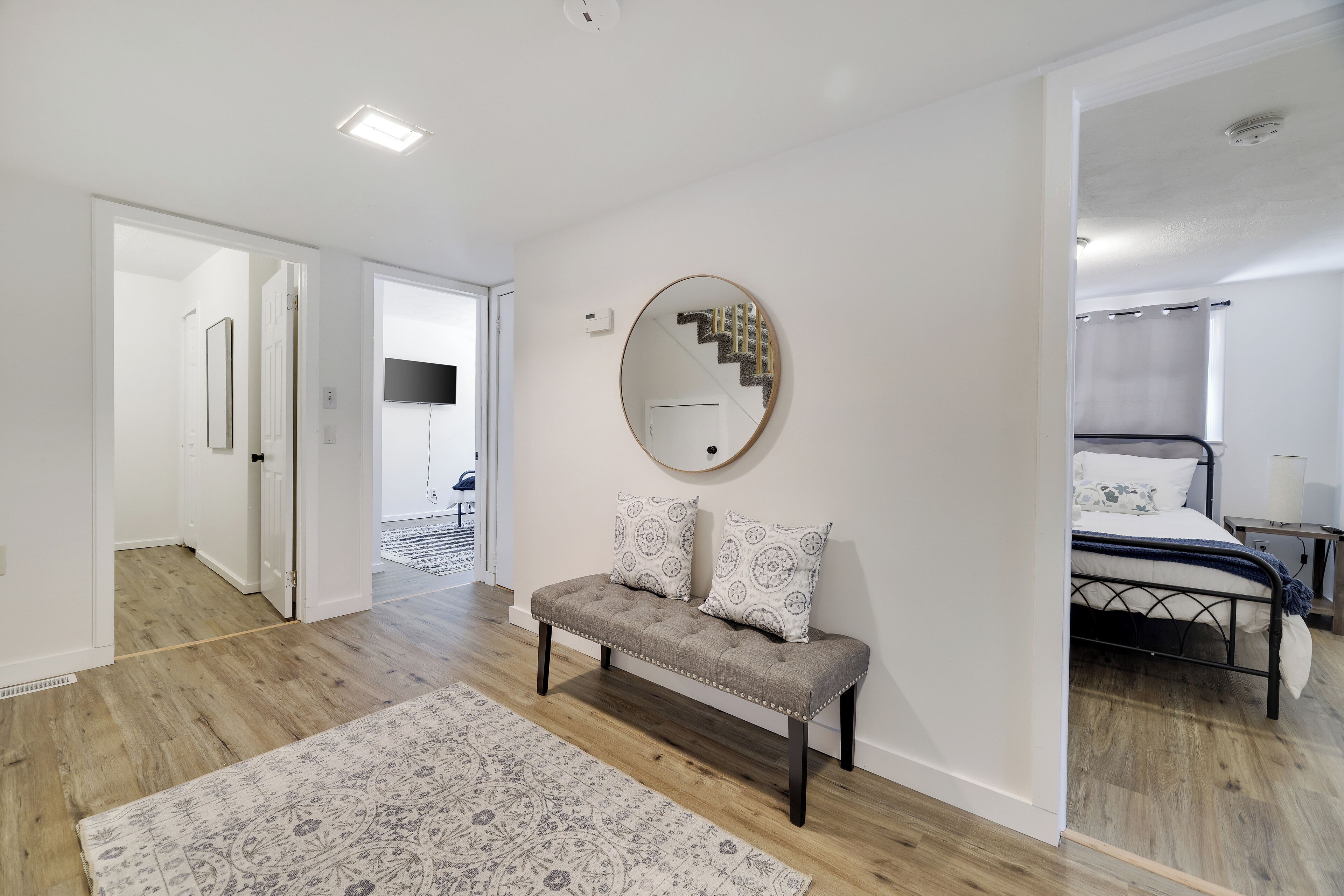 Une chambre moderne avec un grand lit, un banc agrémenté de coussins décoratifs, un miroir rond et un escalier visible au fond.