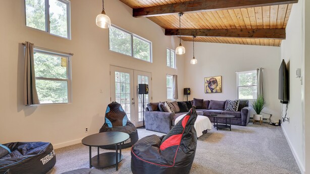 Ein geräumiger Wohnbereich mit großen Fenstern, einer hohen Holzbalkendecke und moderner Möblierung, darunter Sitzsäcke und eine Couch.