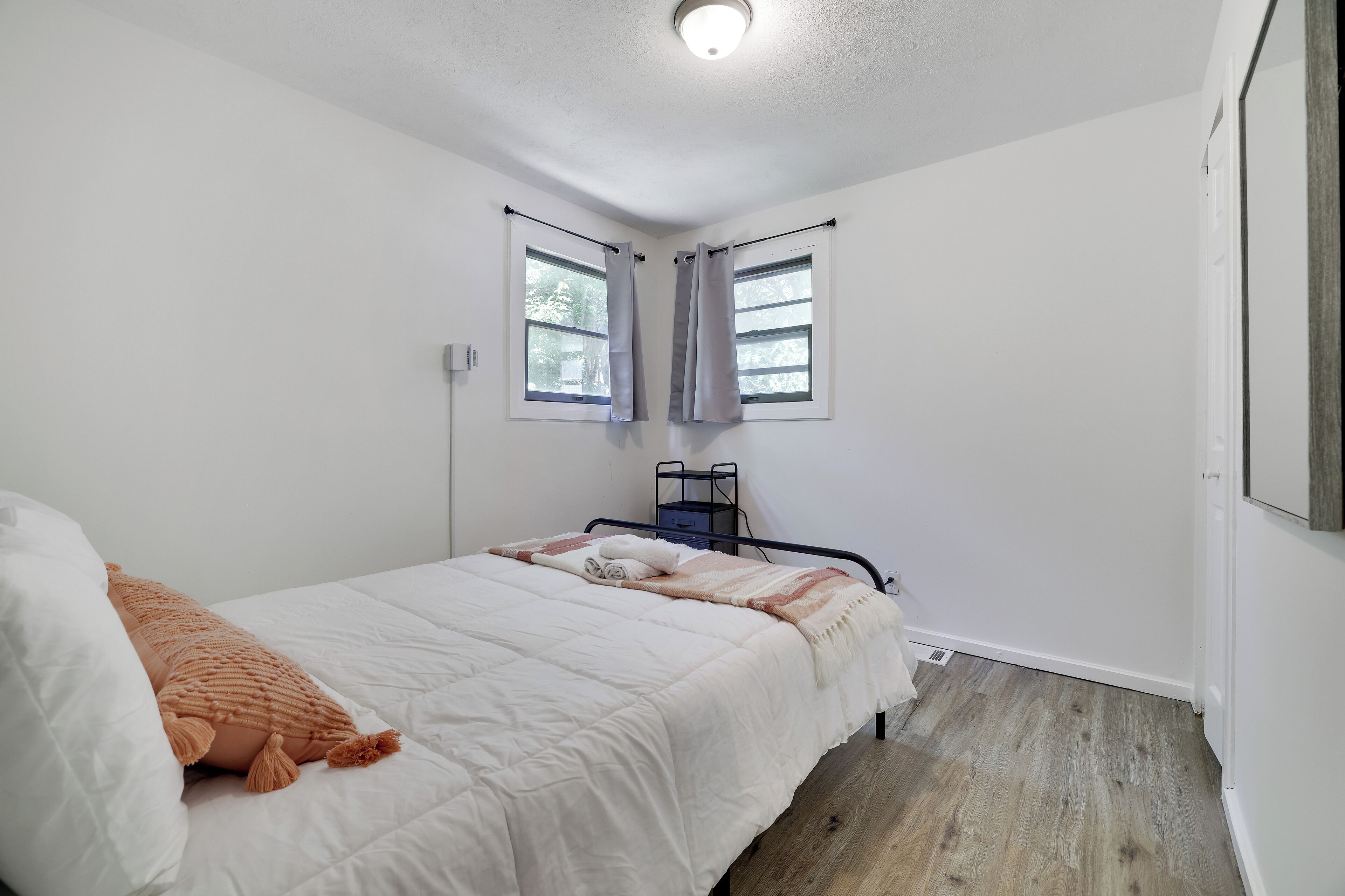 Une chambre à coucher comprenant un lit, une table de chevet, deux fenêtres avec des rideaux et un luminaire au plafond.