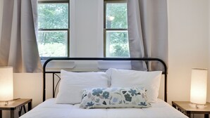 Ein ordentlich bezogenes Bett mit weißer Bettwäsche und einem floralen Kissen, das von zwei Nachttischen mit Lampen begleitet wird, in einem Zimmer mit Fenster und Vorhängen.