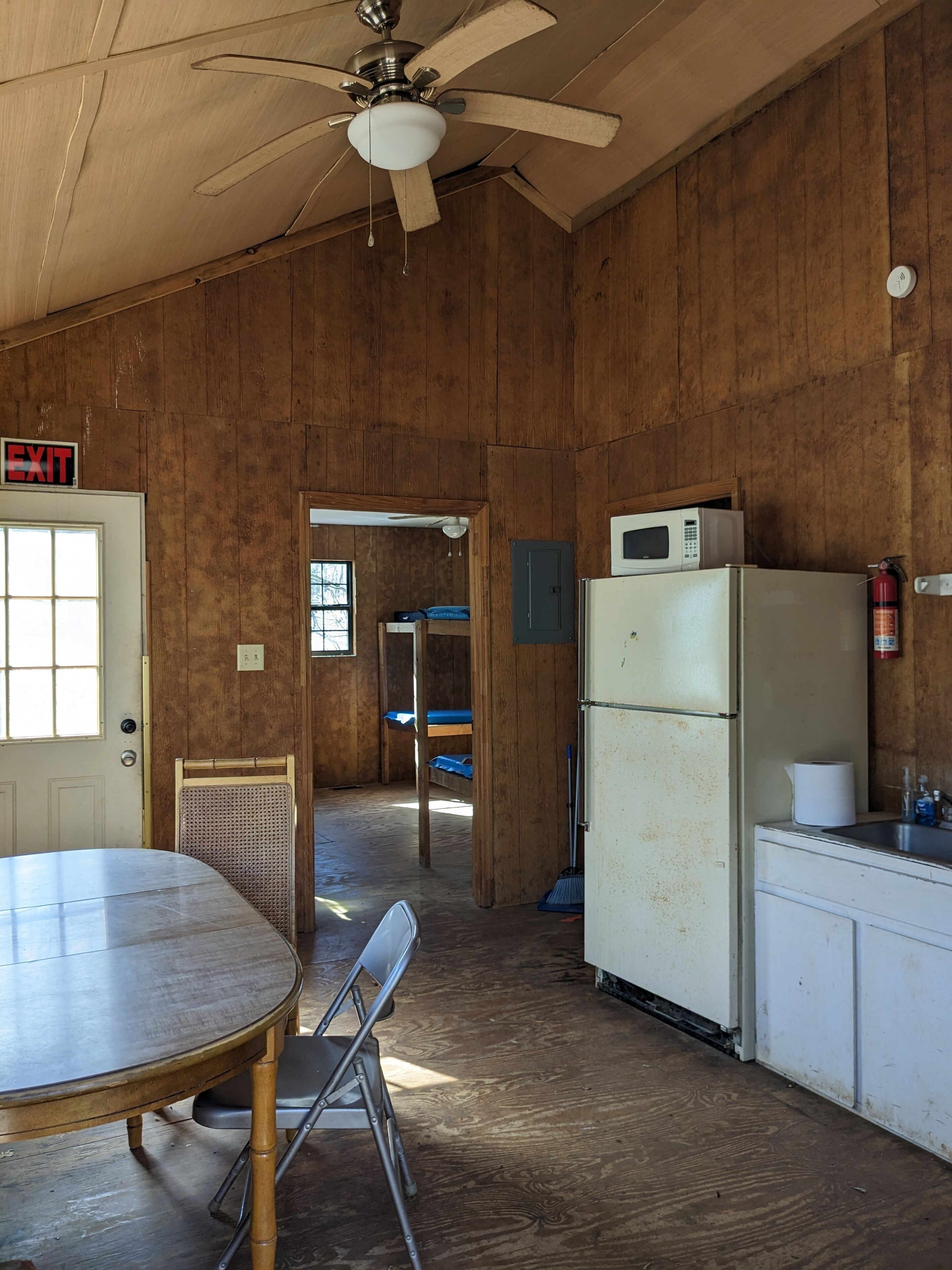 A rustic kitchen with wooden walls, a ceiling fan, a refrigerator, a microwave, and a sink.