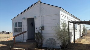 A white single-story building with a front door, a small porch, and a large fan on the side. - Comfy 2 bedroom home, near Palo Duro Canyon! (Canyon)