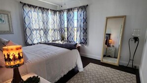 A bedroom with a bed, a nightstand with a lamp, a mirror, and a window with patterned curtains. - Comfy 2 bedroom home, near Palo Duro Canyon! (Canyon)