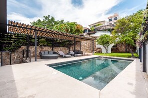 Relax by the clear rectangular pool surrounded by lush greenery and mature trees, with a shaded area beneath the pergola.