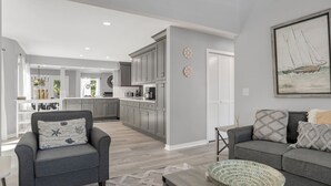 A modern living room with a grey sofa, a grey armchair, and a coffee table. There is a kitchen in the background with grey cabinets and white countertops.