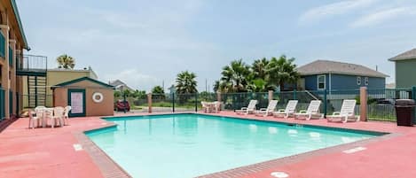A rectangular swimming pool with a red deck, surrounded by lounge chairs and a small table.
