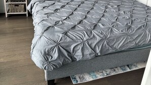 A bedroom with a grey quilted bedspread, a grey upholstered headboard, a bedside table with a lamp, and a wall art piece above the bed.