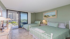 Une chambre avec deux lits, un balcon donnant sur un immeuble de grande hauteur et un tableau accroché au mur.