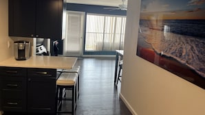 A modern kitchen with dark cabinets, a coffee maker, and a bench. There is a large window with blinds and a painting of a beach scene on the wall. - OCEANFRONT with Balcony, Updated Condo; Gorgeous views; sleeps 6 (Ocean City)