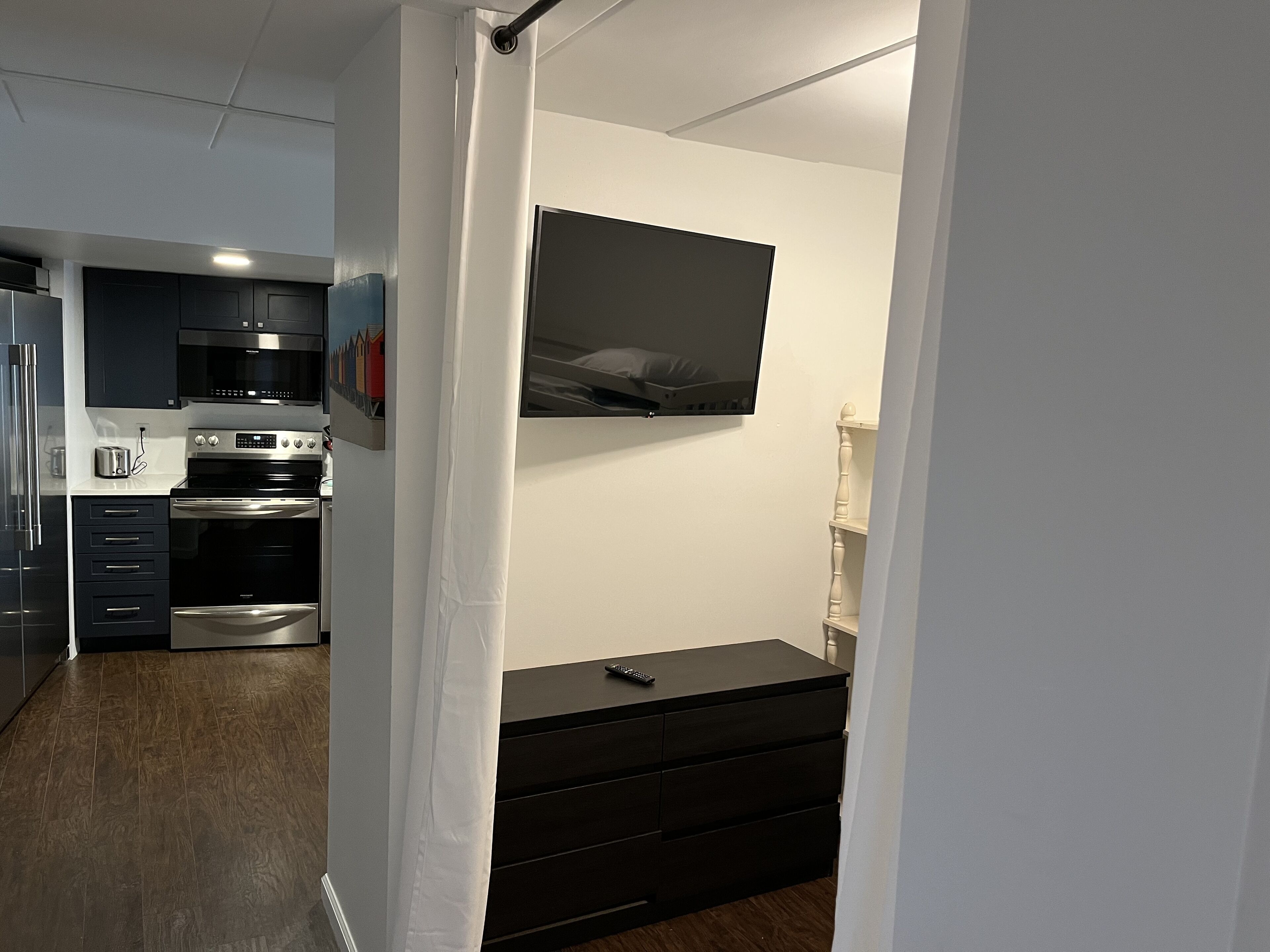 A modern kitchen with dark cabinets, stainless steel appliances, and a flat-screen TV mounted above a black dresser.