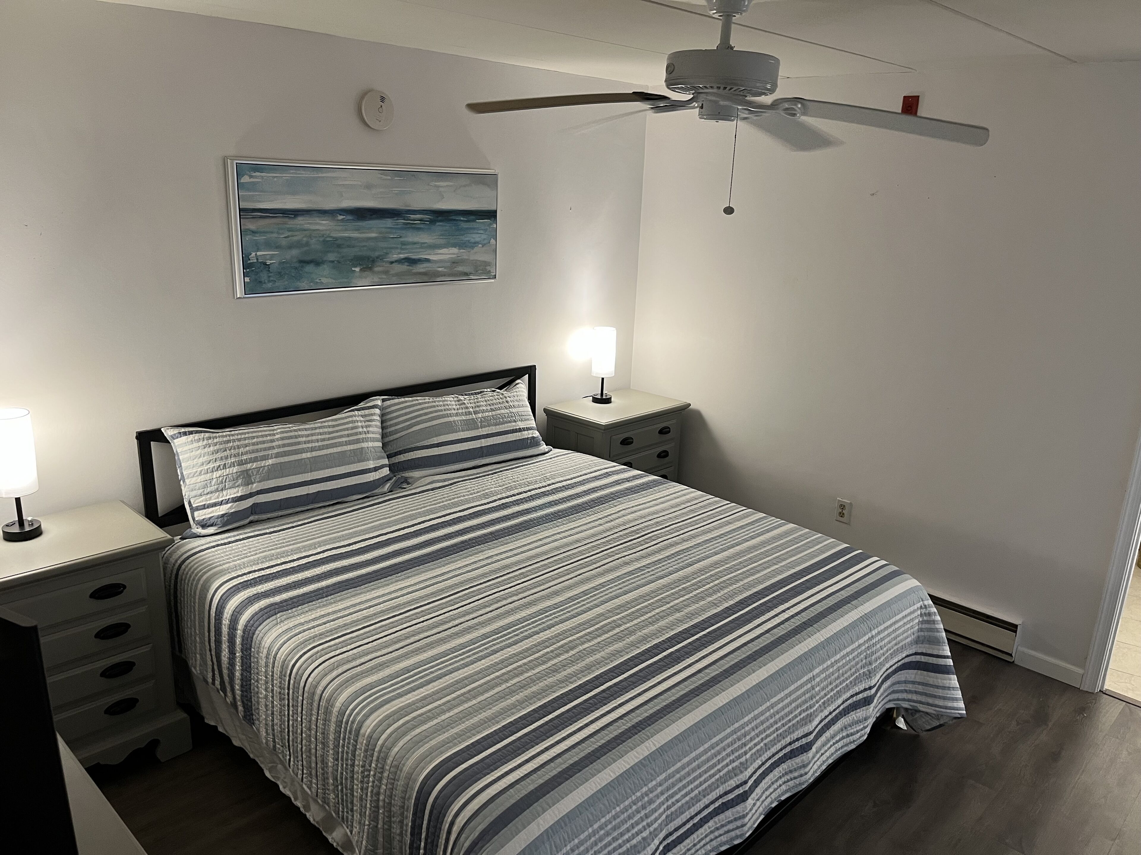 A bedroom with a bed, nightstands, a ceiling fan, a painting, and a wall-mounted light.
