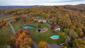 A residential property with a large lawn, a tennis court, a swimming pool, and a house surrounded by autumn-colored trees. - Updated Phase 2 Hideaway Valley Condo Nestled in the Woods (Harbor Springs)