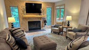 A living room with a fireplace, a flat-screen TV mounted on a wooden wall, and two windows with views of greenery. - Updated Phase 2 Hideaway Valley Condo Nestled in the Woods (Harbor Springs)