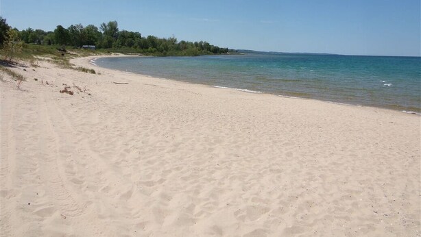 A sandy beach with clear water and a forested area in the background. - Updated Phase 2 Hideaway Valley Condo Nestled in the Woods (Harbor Springs)