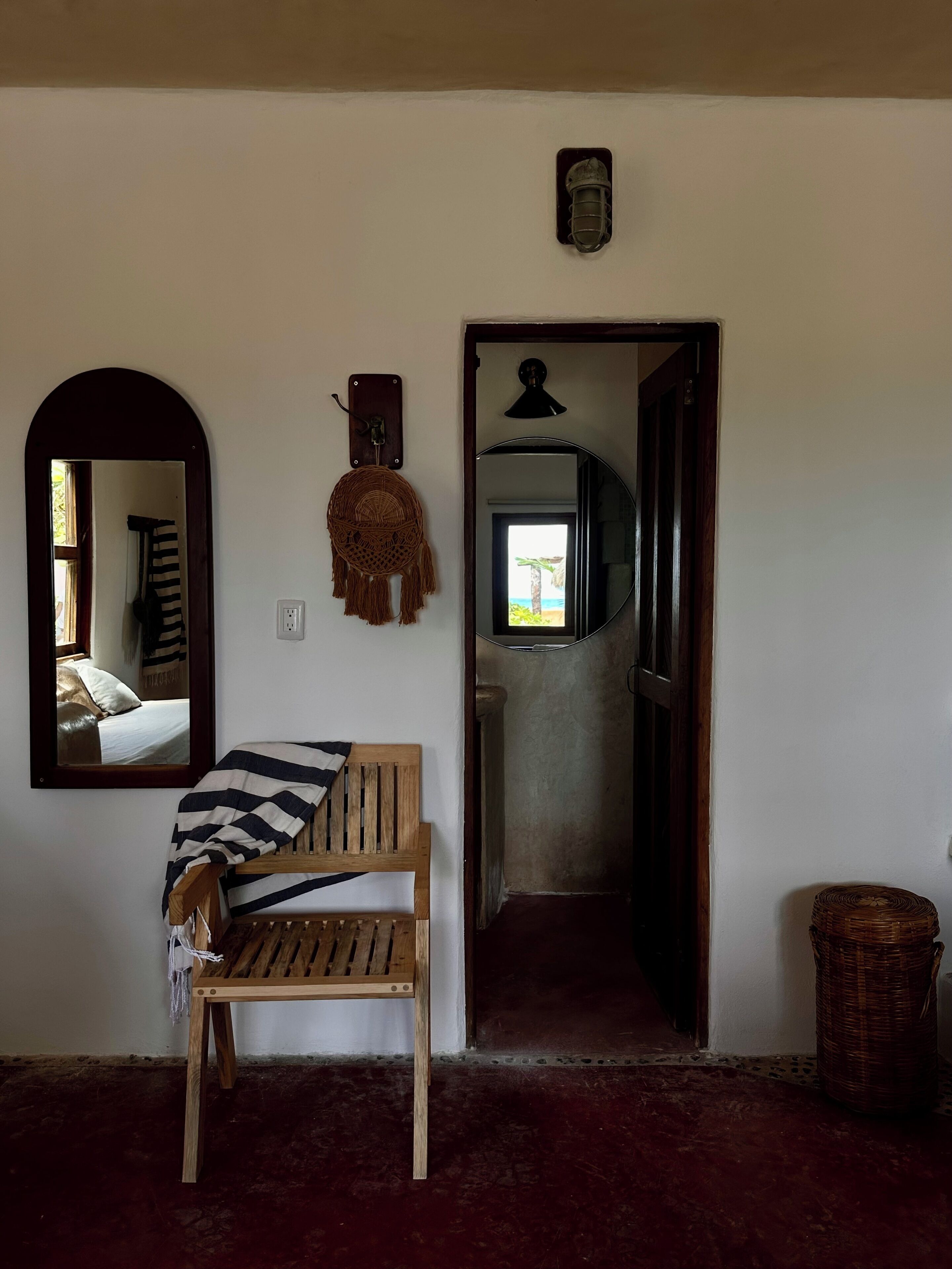 A room with a wooden bench, a mirror, a woven wall hanging, and a doorway leading to another room.