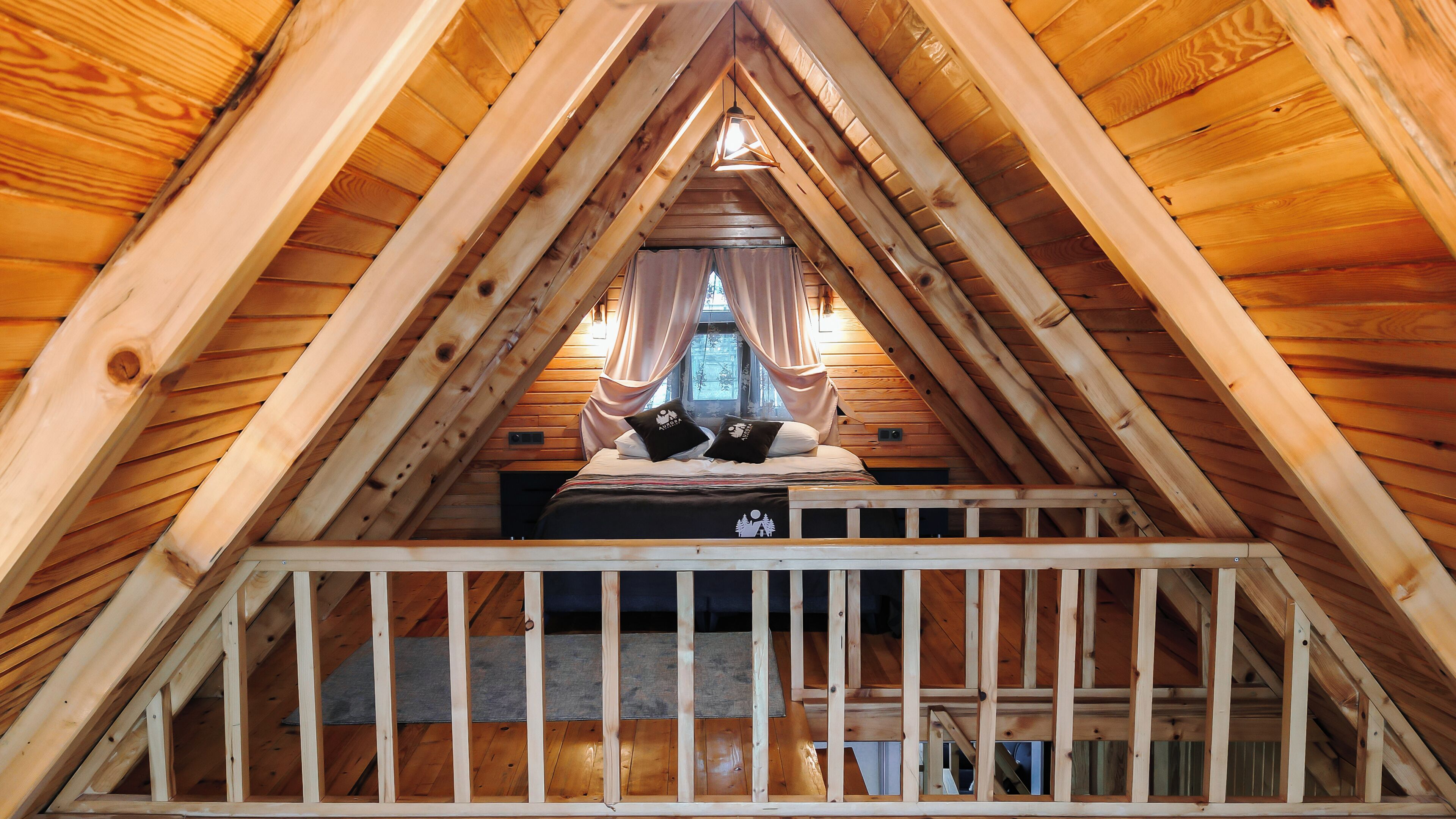 A high-ceilinged attic bedroom with a wooden sloped roof, a bed with two pillows, and a view of the outside through a window.